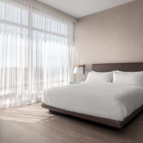 A modern bedroom with a bed, two lamps, TV, sheer curtains on large windows, and wooden flooring, creating a bright, cozy ambiance.
