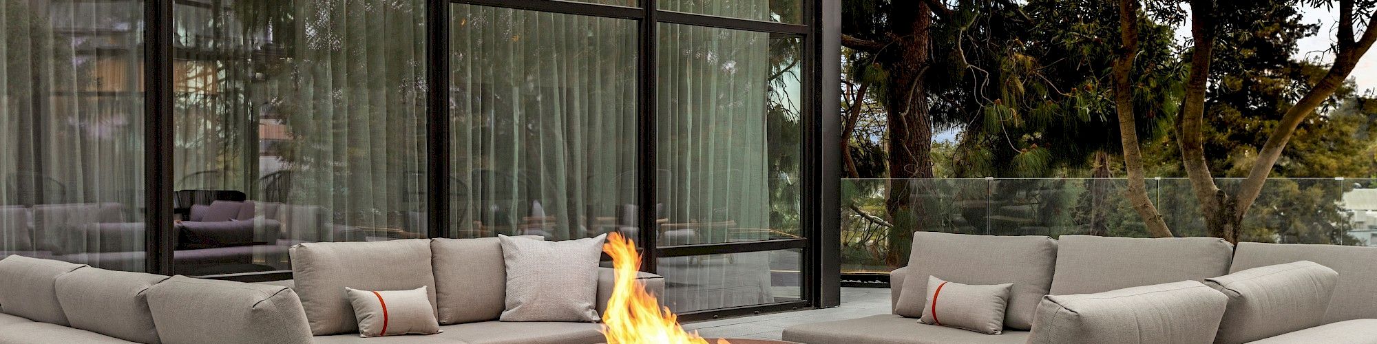 A modern outdoor patio features three beige sofas around a central fire pit, near a building with large glass windows and surrounded by trees.