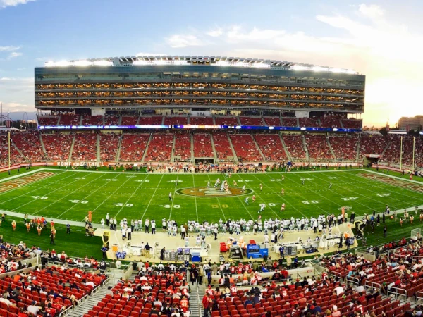 A packed football stadium with teams lined up on the field at dusk, bright lights glowing in the stands and sky.