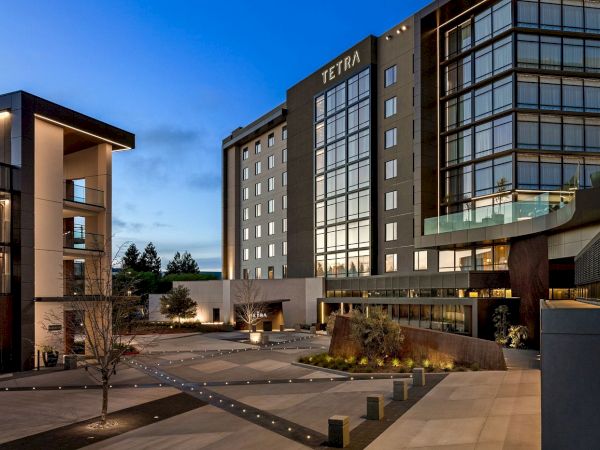 The image shows a modern hotel building with the name "Tetra" on it, featuring large windows, an open courtyard, and evening lighting.