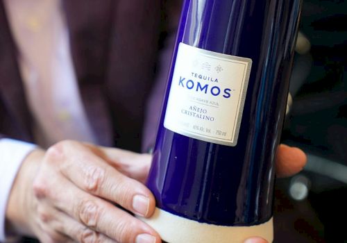 A person holds a blue bottle labeled "Tequila Komos" with a white label, featuring "Añejo Cristalino" and other details.