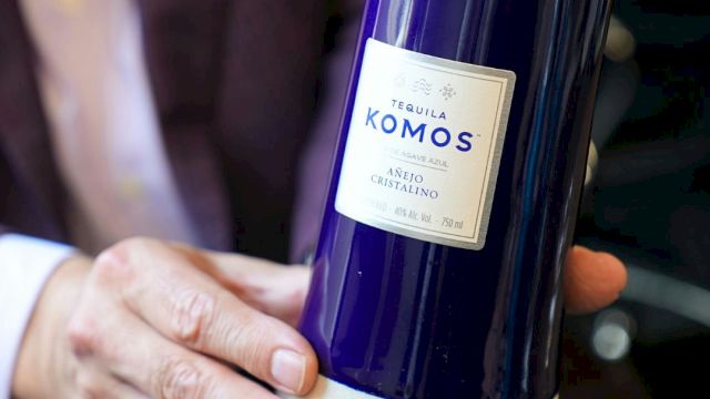 A person holds a blue bottle labeled "Tequila Komos" with a white label, featuring "Añejo Cristalino" and other details.