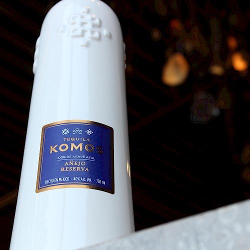 This image shows a white bottle labeled "Tequila Komos Añejo Cristalino" against a blurred background of lights.