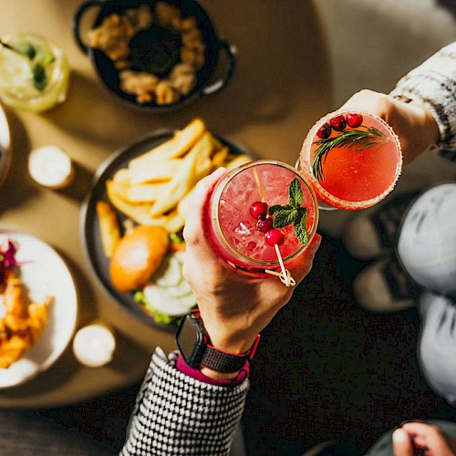 People toasting with red cocktails over a table filled with fries, sliders, and other small dishes, creating a cozy and fun atmosphere.