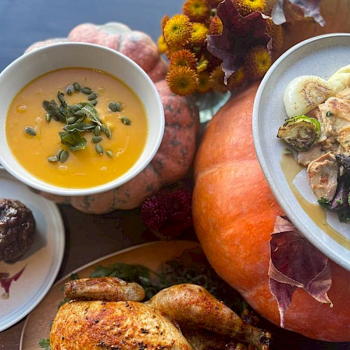 A festive spread with roasted chicken, squash soup, veggies, a dessert, and pumpkins, all on a dark surface with autumn leaves and flowers.