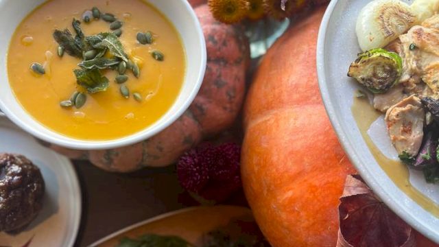 A festive spread with roasted chicken, squash soup, veggies, a dessert, and pumpkins, all on a dark surface with autumn leaves and flowers.
