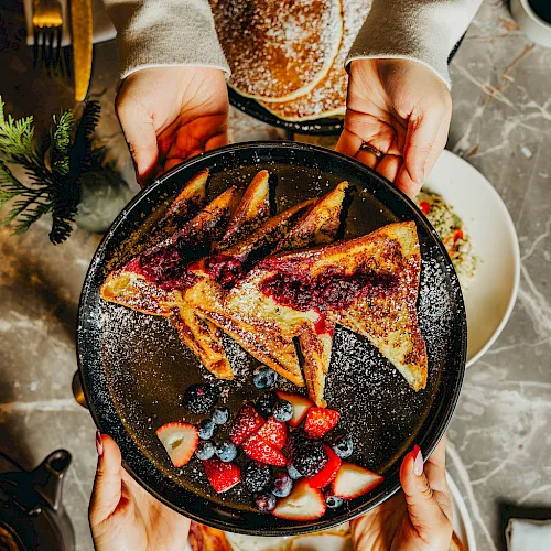 Hands holding a black skillet of golden pancakes topped with berries and powdered sugar, with festive food on the table.