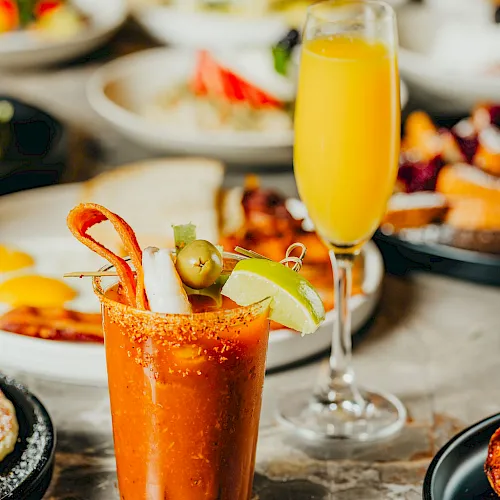 A bar table with an orange cocktail in a garnished glass and a tall flute of juice, amid plated food.