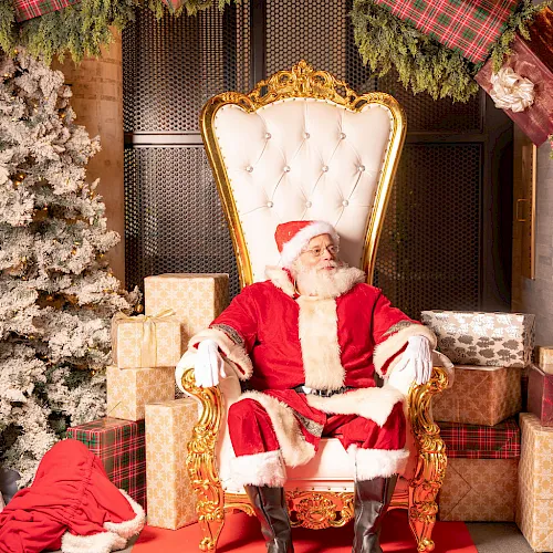 Santa sits in a gold throne in a Christmas display, surrounded by wrapped gifts, evergreen garlands, and a red carpet.