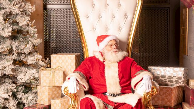 Santa sits in a gold throne in a Christmas display, surrounded by wrapped gifts, evergreen garlands, and a red carpet.
