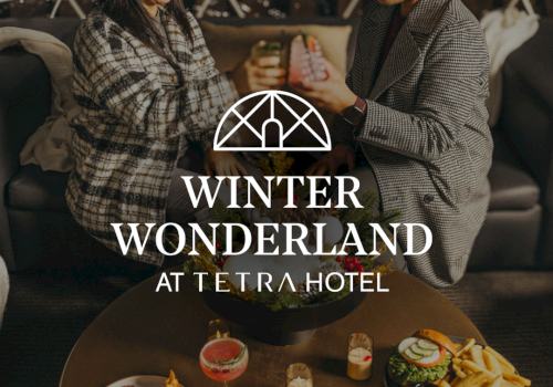 Two people toast in a cozy hotel lounge with a table of desserts and drinks, promoting “Winter Wonderland at Tetra Hotel.”