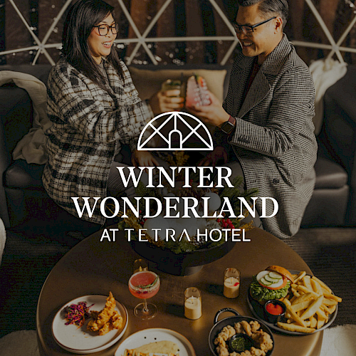 Two people toast in a cozy hotel lounge with a table of desserts and drinks, promoting “Winter Wonderland at Tetra Hotel.”