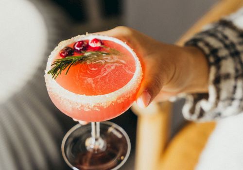 A hand holding a pink grapefruit cocktail in a stemmed glass, garnished with a sprig of rosemary and a small flower in the drink.