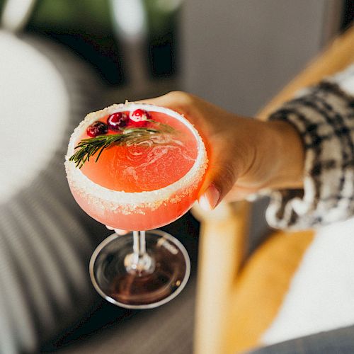 A hand holding a pink grapefruit cocktail in a stemmed glass, garnished with a sprig of rosemary and a small flower in the drink.