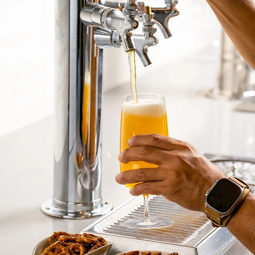 A person pours beer from a tap into a glass, with bowls of pretzels on the counter nearby.