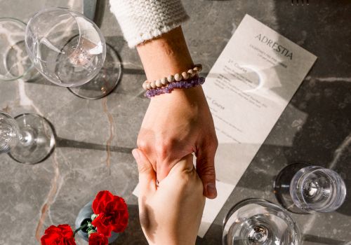 A couple holds hands over a restaurant table, bracelets visible; romantic setting with flowers, glassware, and a shared moment.