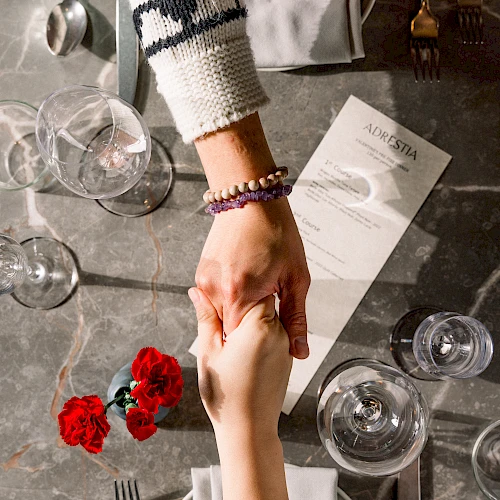 A couple holds hands over a restaurant table, bracelets visible; romantic setting with flowers, glassware, and a shared moment.
