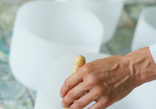 A person uses wooden mallets to strike white singing bowls arranged in a line, likely for sound therapy or meditation.
