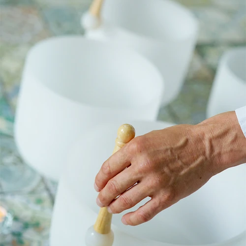 A person uses wooden mallets to strike white singing bowls arranged in a line, likely for sound therapy or meditation.