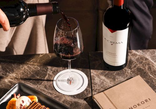 Someone pours red wine as a chocolate dessert and fruit plate awaits, with a bottle and a book on a marble table.