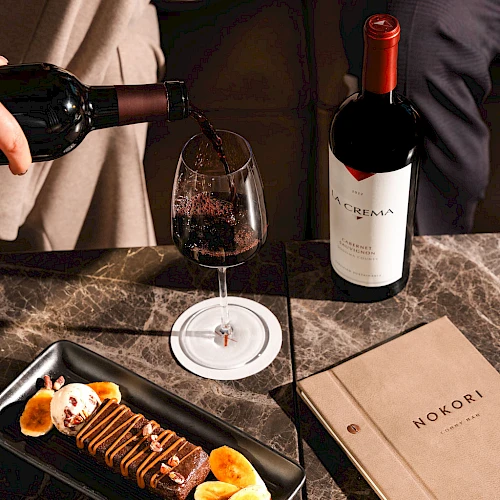 Someone pours red wine as a chocolate dessert and fruit plate awaits, with a bottle and a book on a marble table.