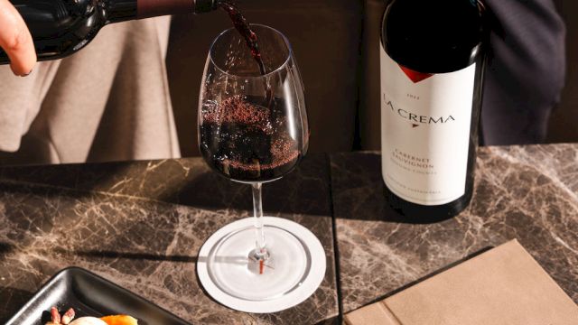 Someone pours red wine as a chocolate dessert and fruit plate awaits, with a bottle and a book on a marble table.
