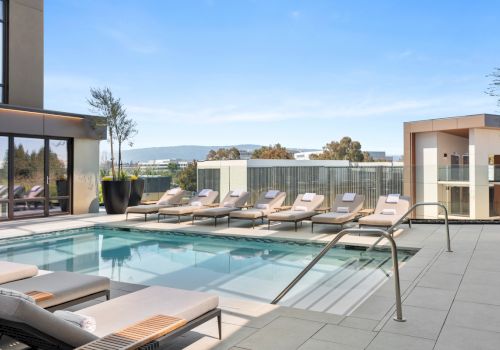 A rooftop pool area with lounge chairs, clear blue water, and glass railings overlooking a city view in the distance.