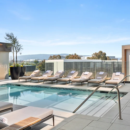 A rooftop pool area with lounge chairs, clear blue water, and glass railings overlooking a city view in the distance.