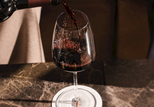 Pouring red wine into a glass placed on a marble counter, a bottle tilted as wine flows smoothly into the goblet.