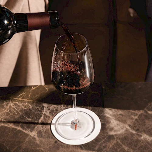 Pouring red wine into a glass placed on a marble counter, a bottle tilted as wine flows smoothly into the goblet.