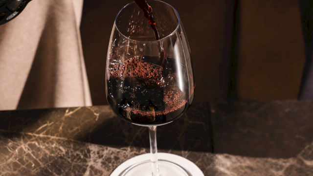 Pouring red wine into a glass placed on a marble counter, a bottle tilted as wine flows smoothly into the goblet.