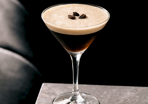 A stylish cocktail in a stemmed glass with a frothy top and coffee beans garnish, set against a dark, moody backdrop.