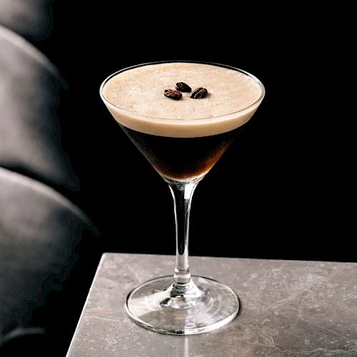 A stylish cocktail in a stemmed glass with a frothy top and coffee beans garnish, set against a dark, moody backdrop.