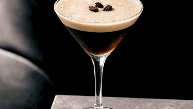 A stylish cocktail in a stemmed glass with a frothy top and coffee beans garnish, set against a dark, moody backdrop.