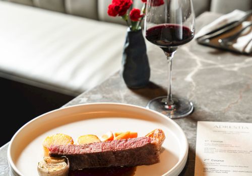 A fine dining table with a plated dessert, red wine, and a small vase of red flowers on a marble counter, set against a plush booth.