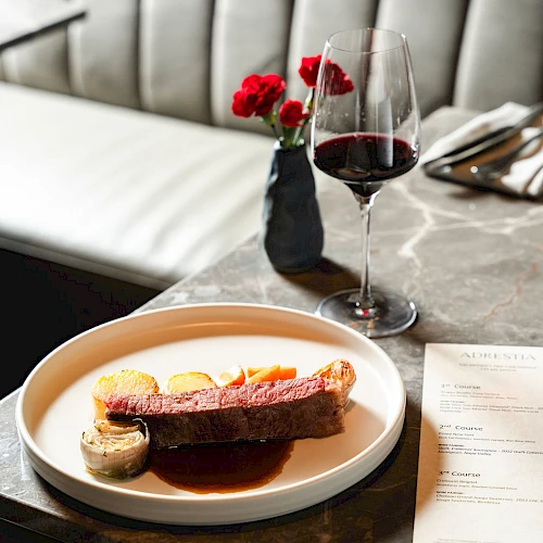 A fine dining table with a plated dessert, red wine, and a small vase of red flowers on a marble counter, set against a plush booth.