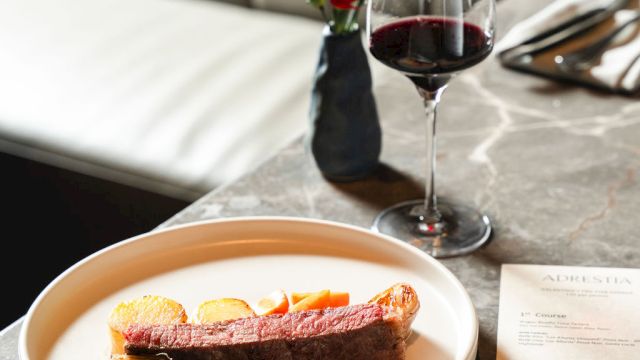 A fine dining table with a plated dessert, red wine, and a small vase of red flowers on a marble counter, set against a plush booth.