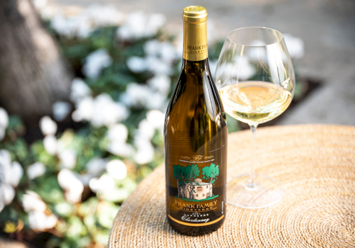 A bottle of white wine with a glass on a wooden table outdoors, a sunny garden setting.