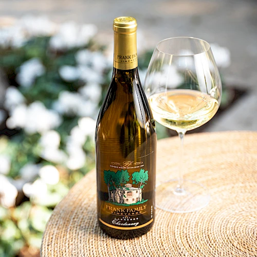 A bottle of white wine with a glass on a wooden table outdoors, a sunny garden setting.