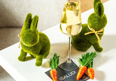 Two mossy bunny decorations toast with a glass of champagne on a white table, surrounded by small carrots for a whimsical setup.