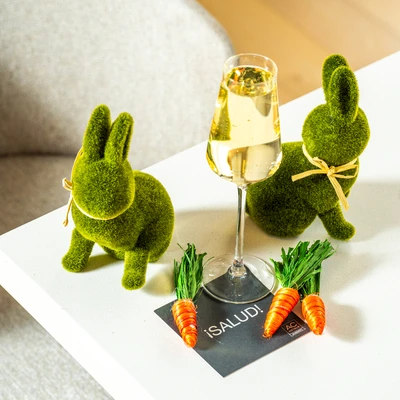 Two mossy bunny decorations toast with a glass of champagne on a white table, surrounded by small carrots for a whimsical setup.