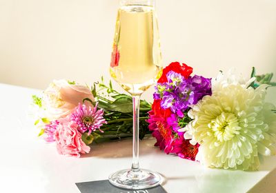 A flute of champagne with a colorful bouquet in the background, resting on a white table near a business card.