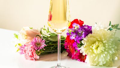 A flute of champagne with a colorful bouquet in the background, resting on a white table near a business card.