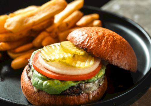 A juicy hamburger with lettuce, tomato, onion, pickles, cheese, and a fried egg on a toasted bun, served with crispy french fries alongside.