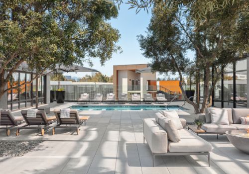 A modern outdoor patio with a rectangular pool, sun loungers, sofa seating, trees, and a contemporary building at the far end, sunny and inviting.