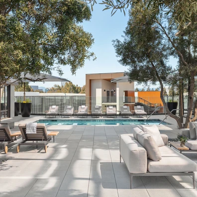 A modern outdoor patio with a rectangular pool, sun loungers, sofa seating, trees, and a contemporary building at the far end, sunny and inviting.