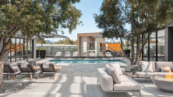 A modern outdoor patio with a rectangular pool, sun loungers, sofa seating, trees, and a contemporary building at the far end, sunny and inviting.