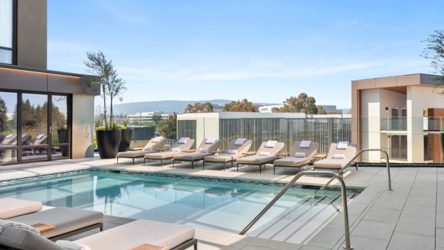 A rooftop pool area with a long rectangular pool, beige lounge chairs along the edge, glass railing, and modern buildings in the background under a clear blue sky.