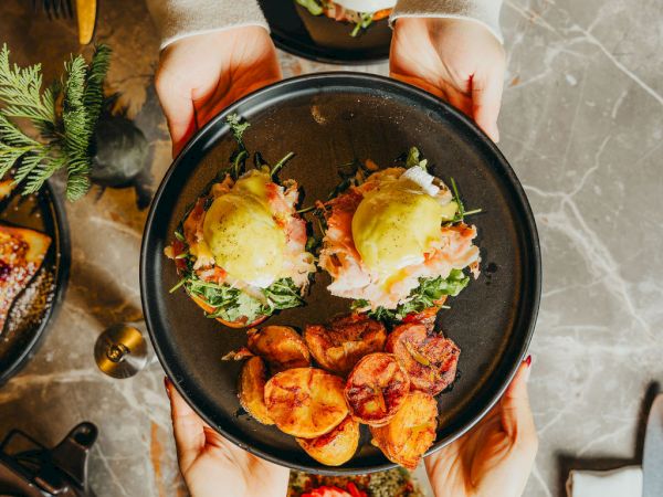 Two hands pass a plate of eggs benedict with fried potatoes to another pair; a sunny-side egg breakfast sits nearby, coffee on the table. End.