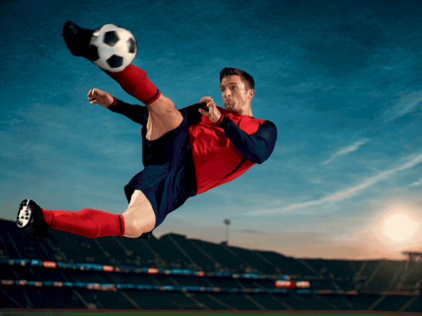 Soccer player in red and blue kit performs a dramatic mid-air kick, stadium, blue sky, and sun on the horizon.